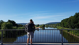 Ein Mann mit Rucksack steht auf einer Brücke und schaut auf einen Fluß